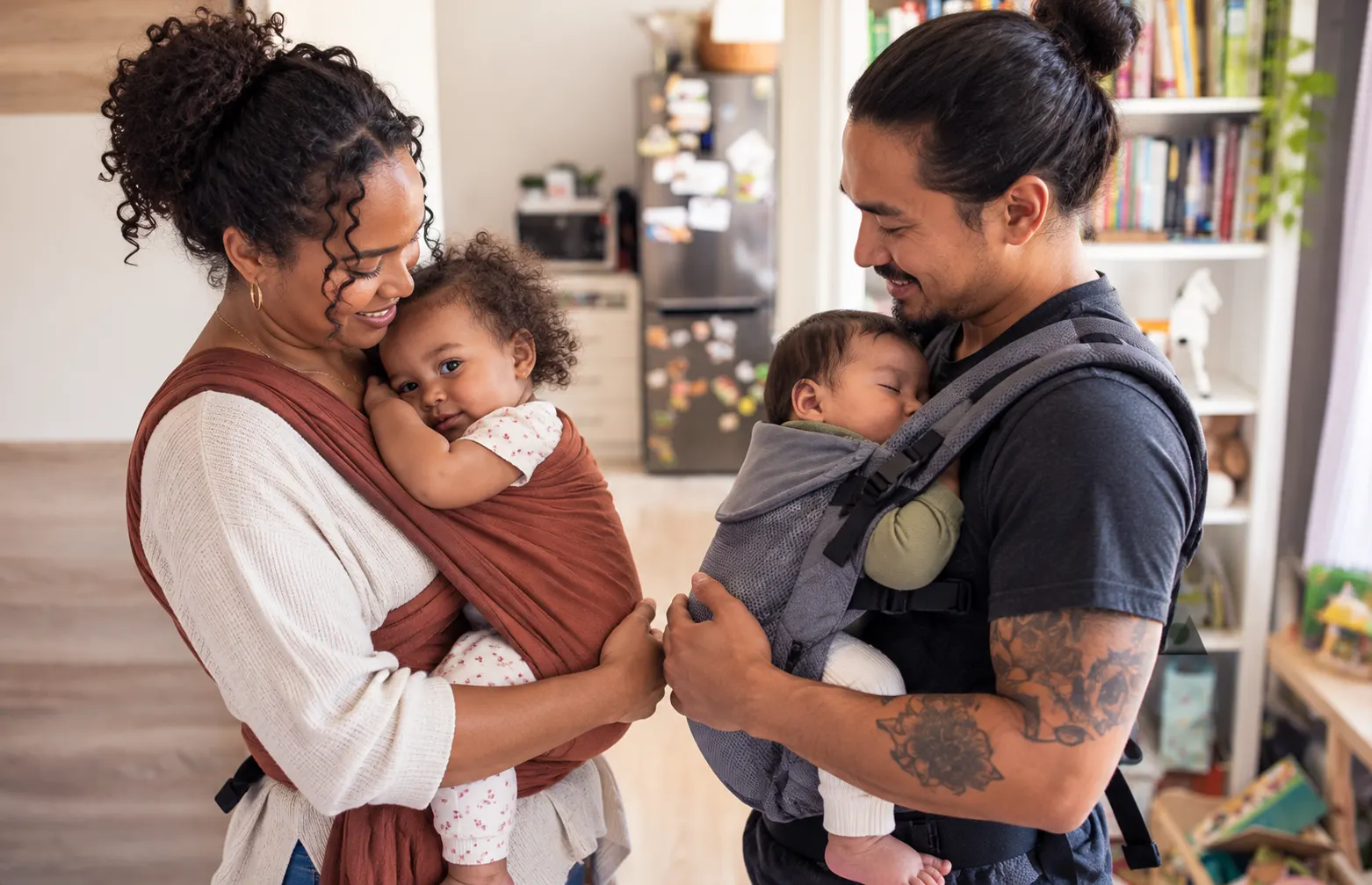 Two parents wearing babies at home: one in a rust-colored woven wrap, the other in a gray soft structured carrier.